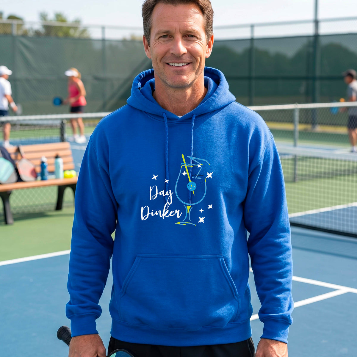 A man wearing a royal blue hoodie with a colorful graphic on the chest that reads 'Day Dinker' alongside a martini cocktail glass with a pickelball olive garnish illustration.