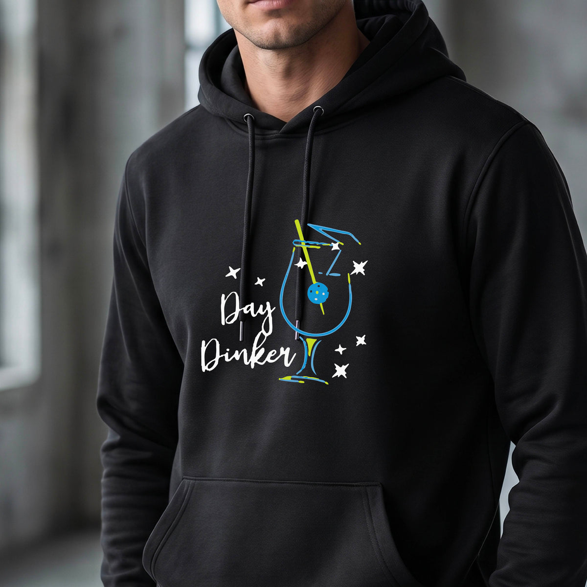 A man wearing a black hoodie with a colorful graphic on the chest that reads 'Day Dinker' alongside a martini cocktail glass with a pickelball olive garnish illustration.