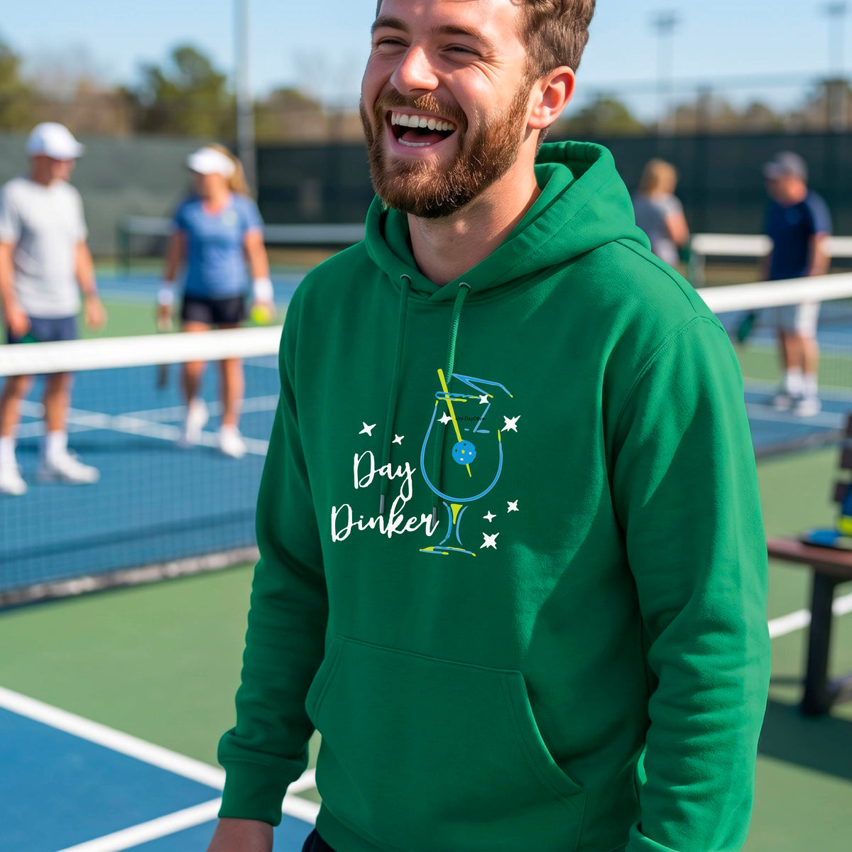 A man wearing a green hoodie with a colorful graphic on the chest that reads 'Day Dinker' alongside a martini cocktail glass with a pickelball olive garnish illustration.
