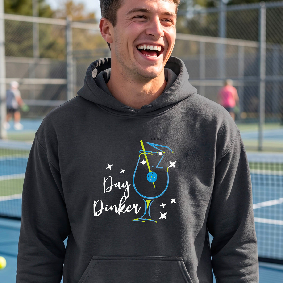 A man wearing a gray hoodie with a colorful graphic on the chest that reads 'Day Dinker' alongside a martini cocktail glass with a pickelball olive garnish illustration.
