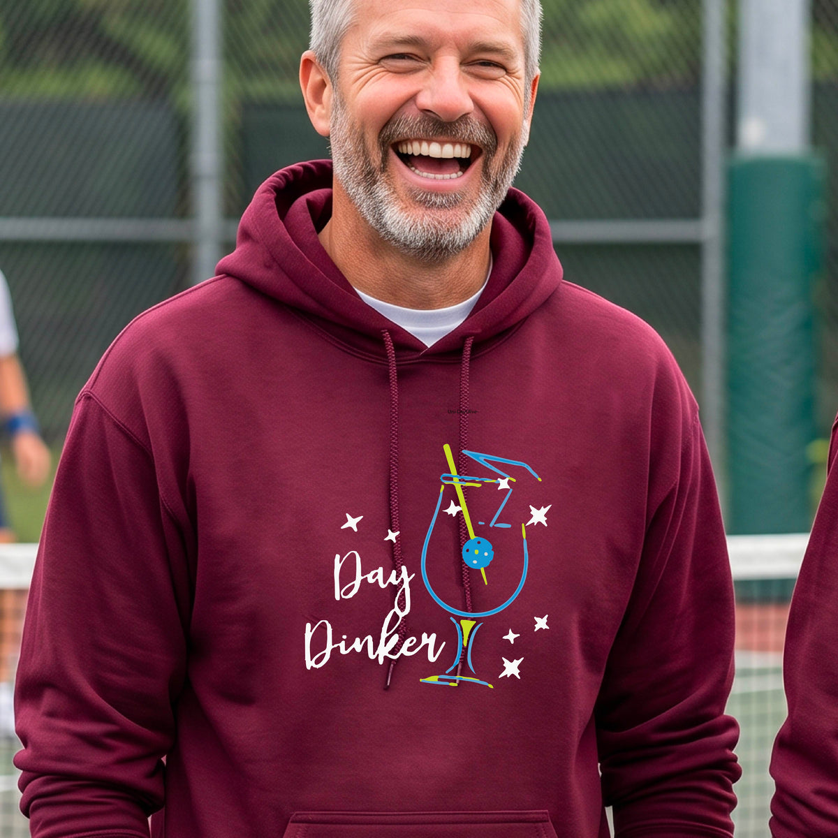 A man wearing a maroon hoodie with a colorful graphic on the chest that reads 'Day Dinker' alongside a martini cocktail glass with a pickelball olive garnish illustration.
