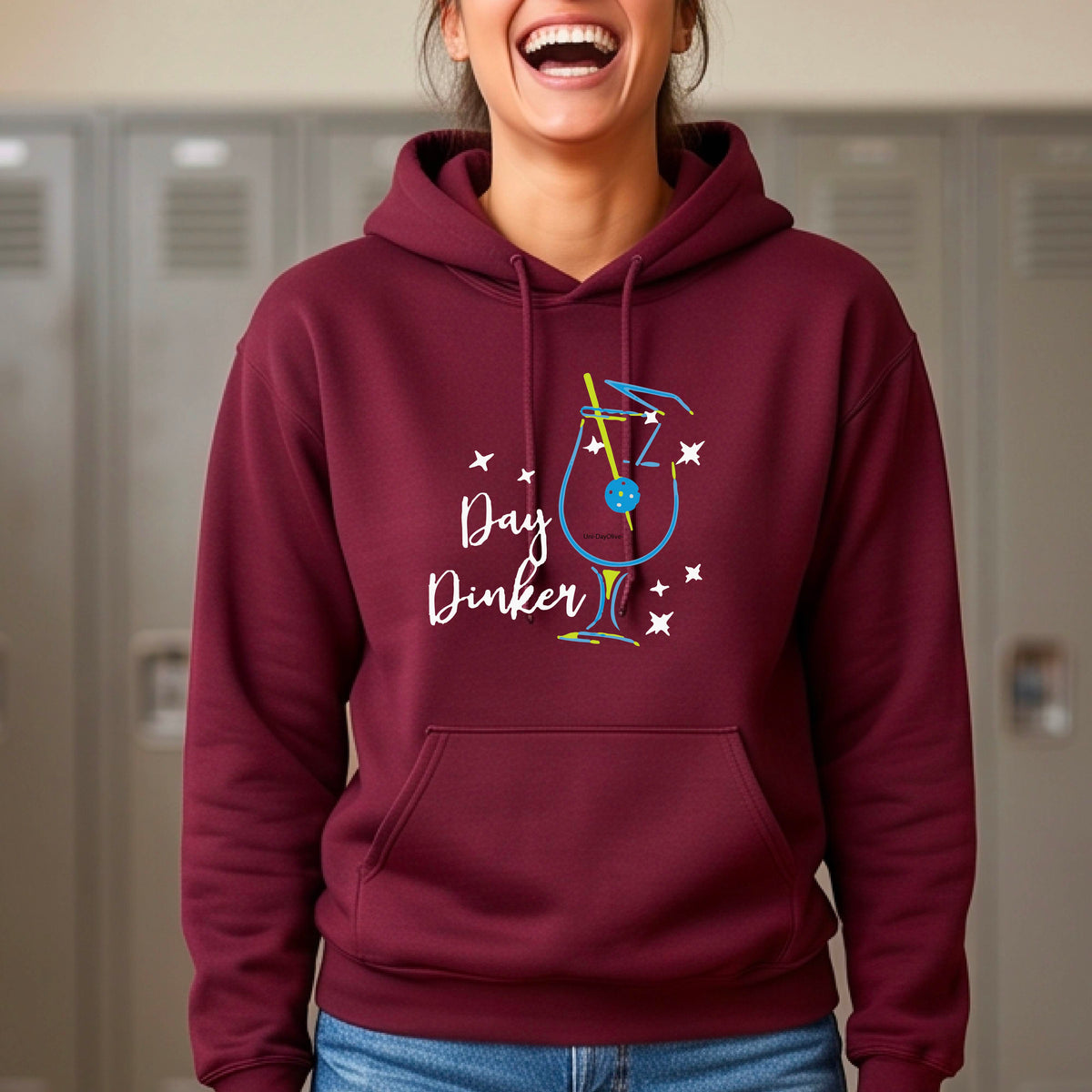 A man wearing a maroon hoodie with a colorful graphic on the chest that reads 'Day Dinker' alongside a martini cocktail glass with a pickelball olive garnish illustration.
