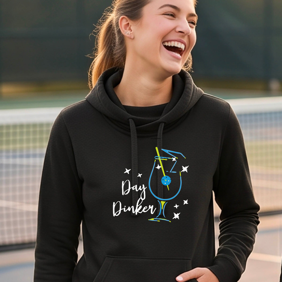A woman wearing a black hoodie with a graphic print that reads 'Day Dinker' in white script, accompanied by a blue and yellow cocktail glass and a pickelball olive garnish illustration and small white stars. 
