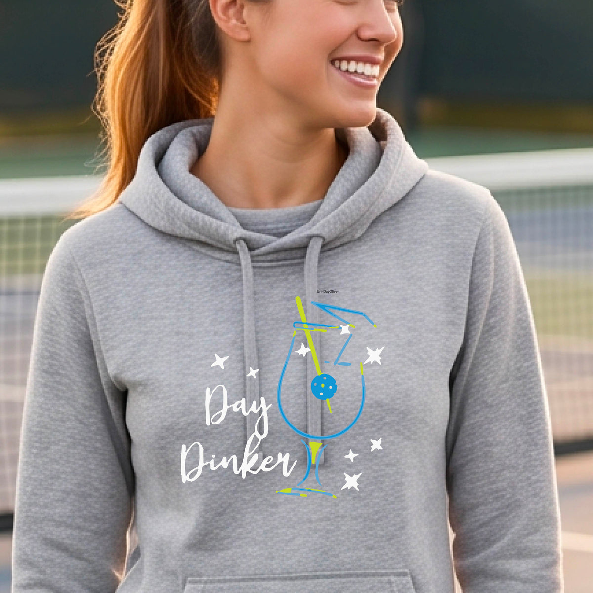 A woman wearing a heather gray hoodie with a graphic print that reads 'Day Dinker' in white script, accompanied by a blue and yellow cocktail glass and a pickelball olive garnish illustration and small white stars. 