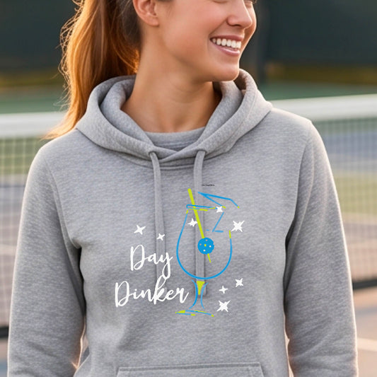 A woman wearing a heather gray hoodie with a graphic print that reads 'Day Dinker' in white script, accompanied by a blue and yellow cocktail glass and a pickelball olive garnish illustration and small white stars. 