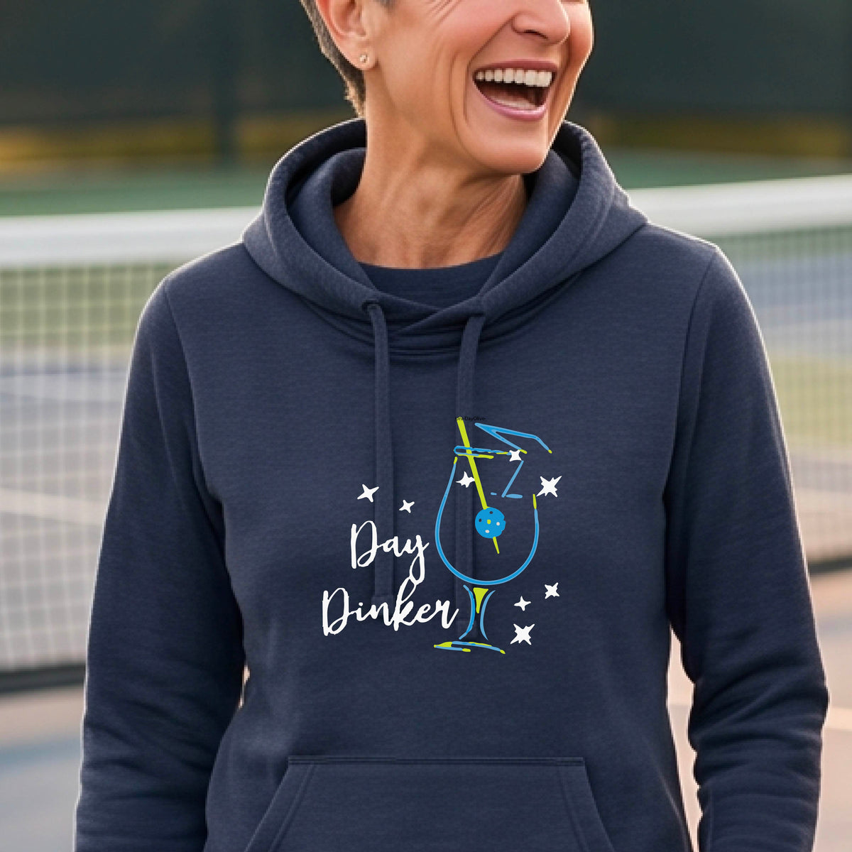 A woman wearing a navy blue hoodie with a graphic print that reads 'Day Dinker' in white script, accompanied by a blue and yellow cocktail glass and a pickelball olive garnish illustration and small white stars. 