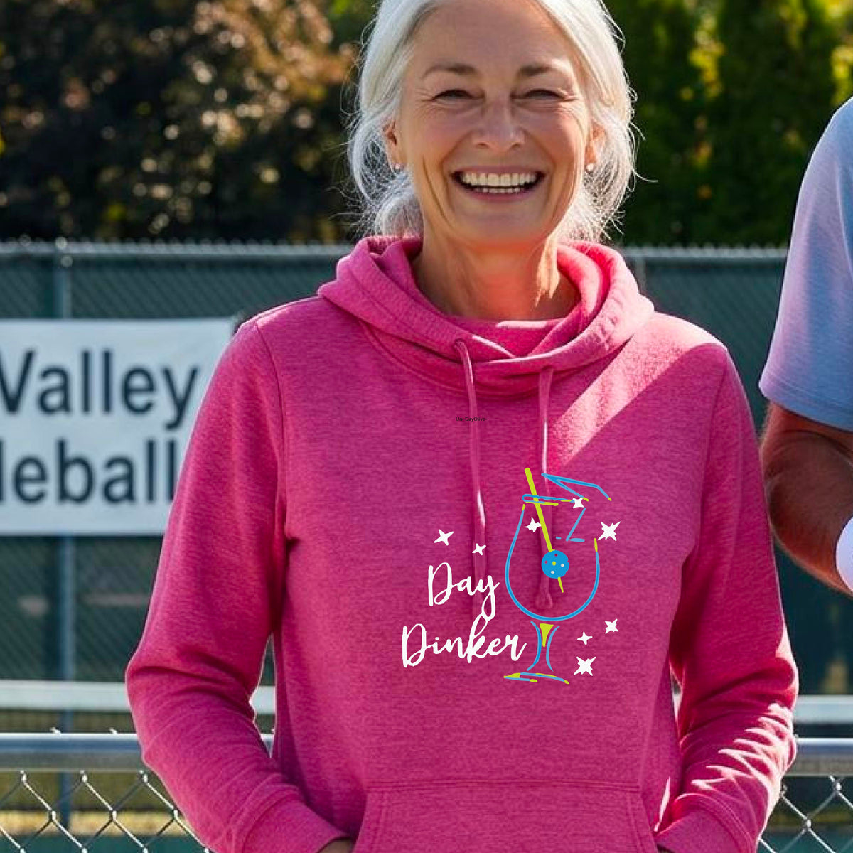 A woman wearing a pink hoodie with a graphic print that reads 'Day Dinker' in white script, accompanied by a blue and yellow cocktail glass and a pickelball olive garnish illustration and small white stars. 