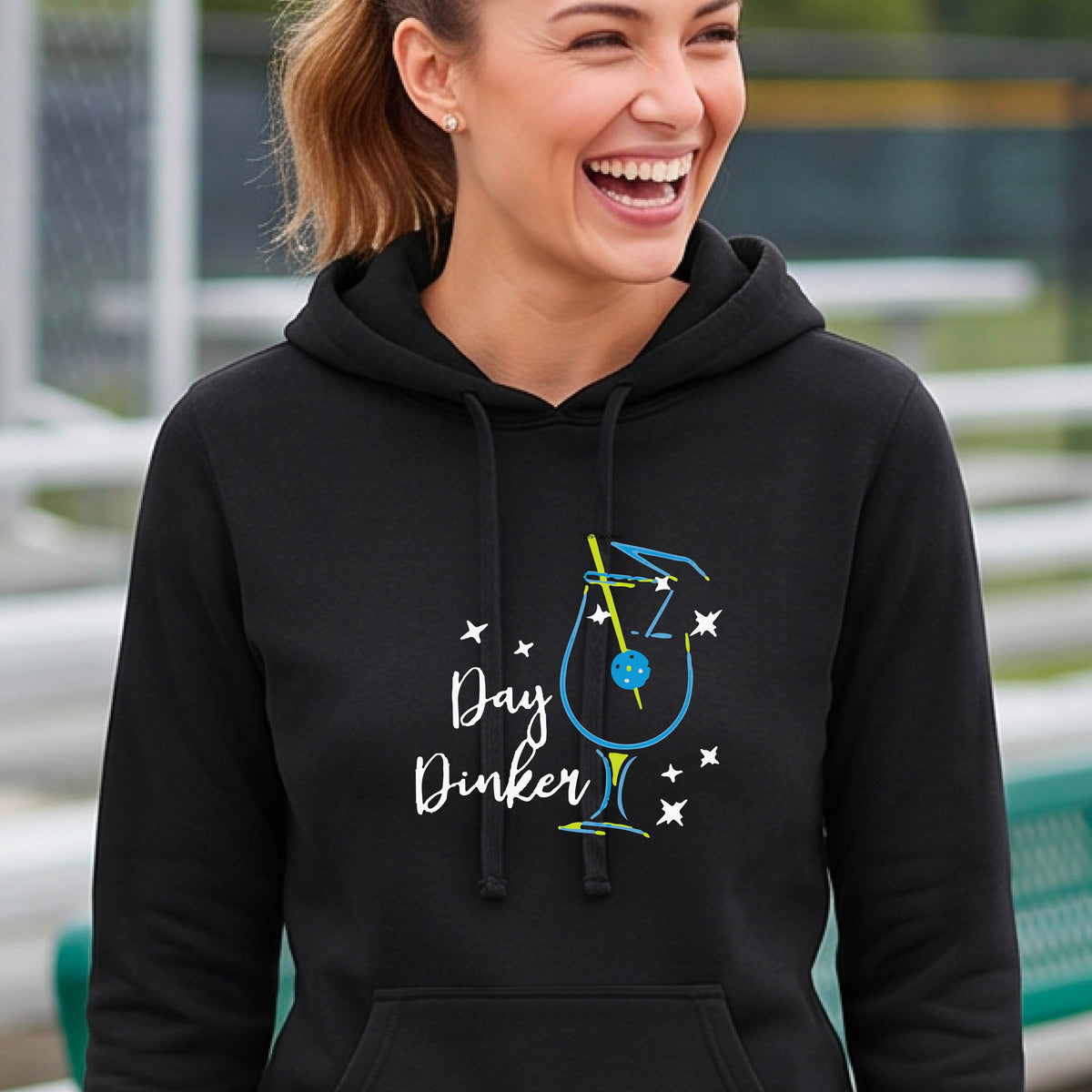 A woman smiling outdoors wearing a lack pullover hoodie with a colorful graphic on the chest that reads 'Day Dinker' with a cocktail illustration and a pickleball olive garnish and small star accents.