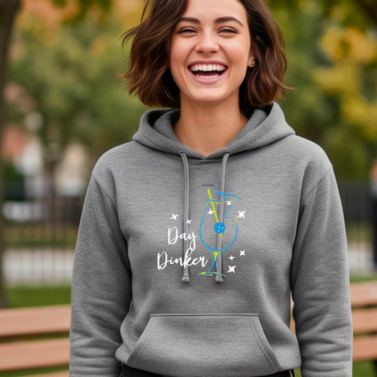 A woman smiling outdoors wearing a heather gray pullover hoodie with a colorful graphic on the chest that reads 'Day Dinker' with a cocktail illustration and a pickleball olive garnish and small star accents.