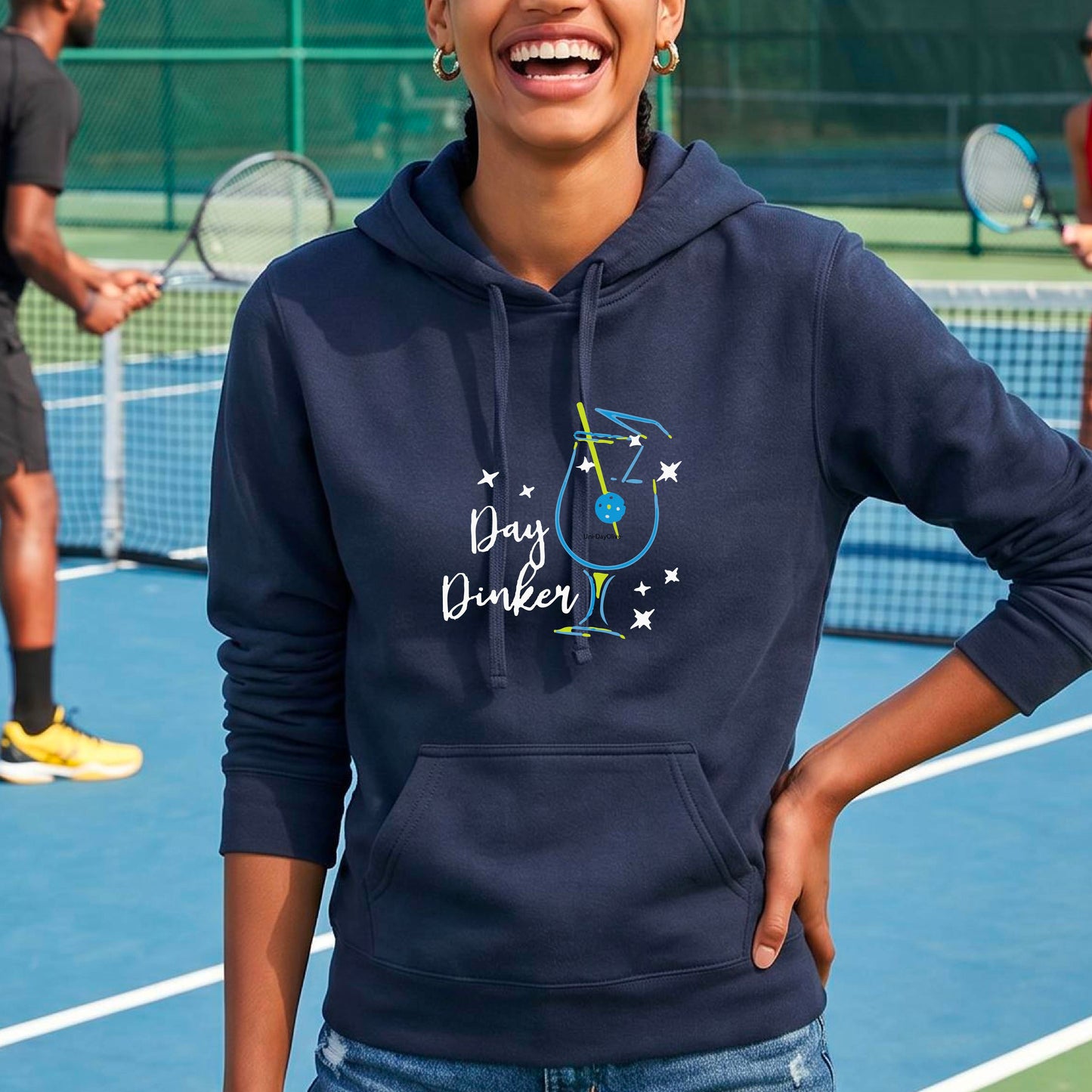A woman smiling outdoors wearing a navy blue pullover hoodie with a colorful graphic on the chest that reads 'Day Dinker' with a cocktail illustration and a pickleball olive garnish and small star accents.