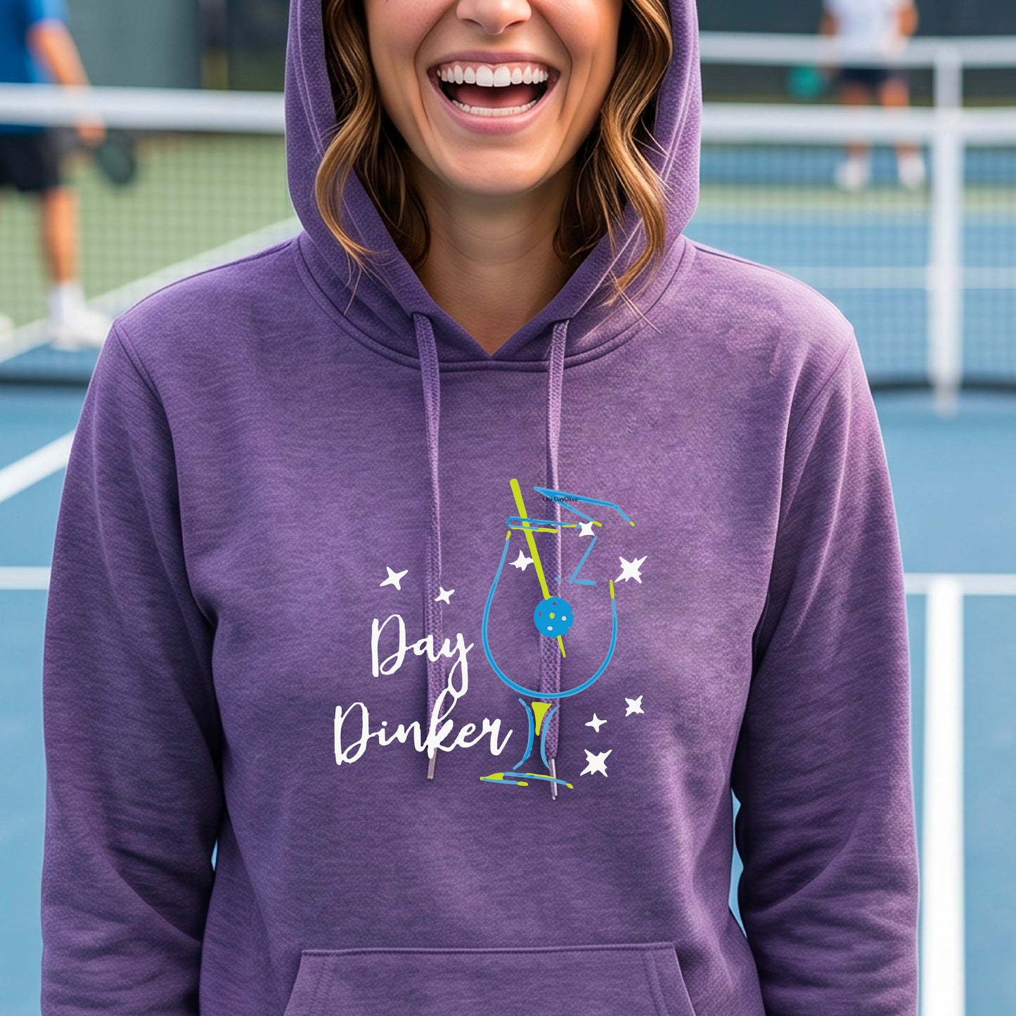 A woman smiling outdoors wearing a purple pullover hoodie with a colorful graphic on the chest that reads 'Day Dinker' with a cocktail illustration and a pickleball olive garnish and small star accents.