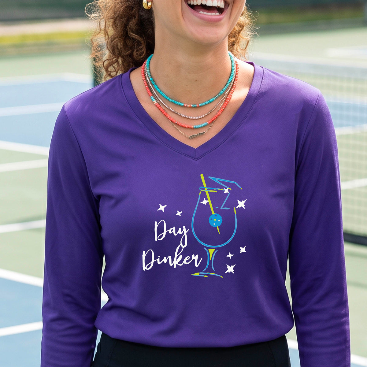 A woman wearing a purple long-sleeve V-neck t-shirt with a 'Day Dinker' cocktail martini graphic on the front, with a pickleball olive garnish.