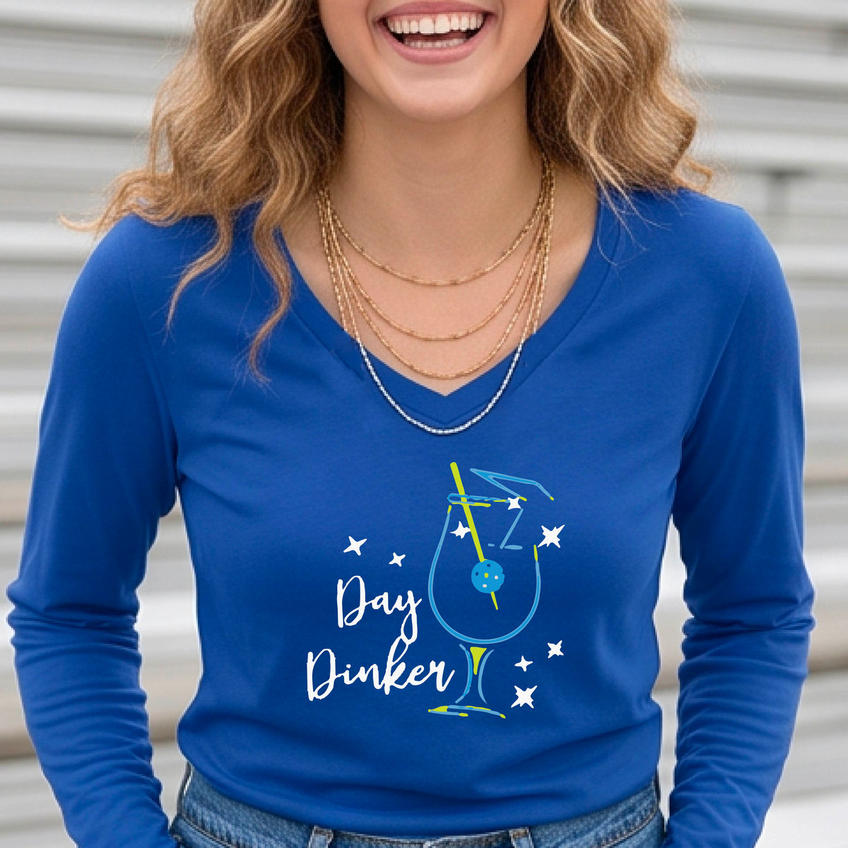 A woman wearing a royal blue long-sleeve V-neck t-shirt with a 'Day Dinker' cocktail martini graphic on the front, with a pickleball olive garnish.