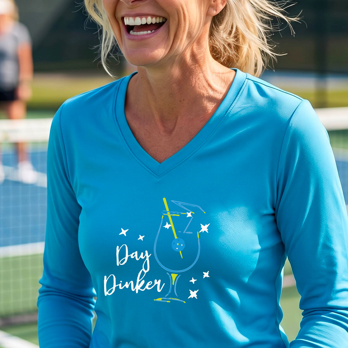 A woman wearing a light blue long-sleeve V-neck t-shirt with a 'Day Dinker' cocktail martini graphic on the front, with a pickleball olive garnish.
