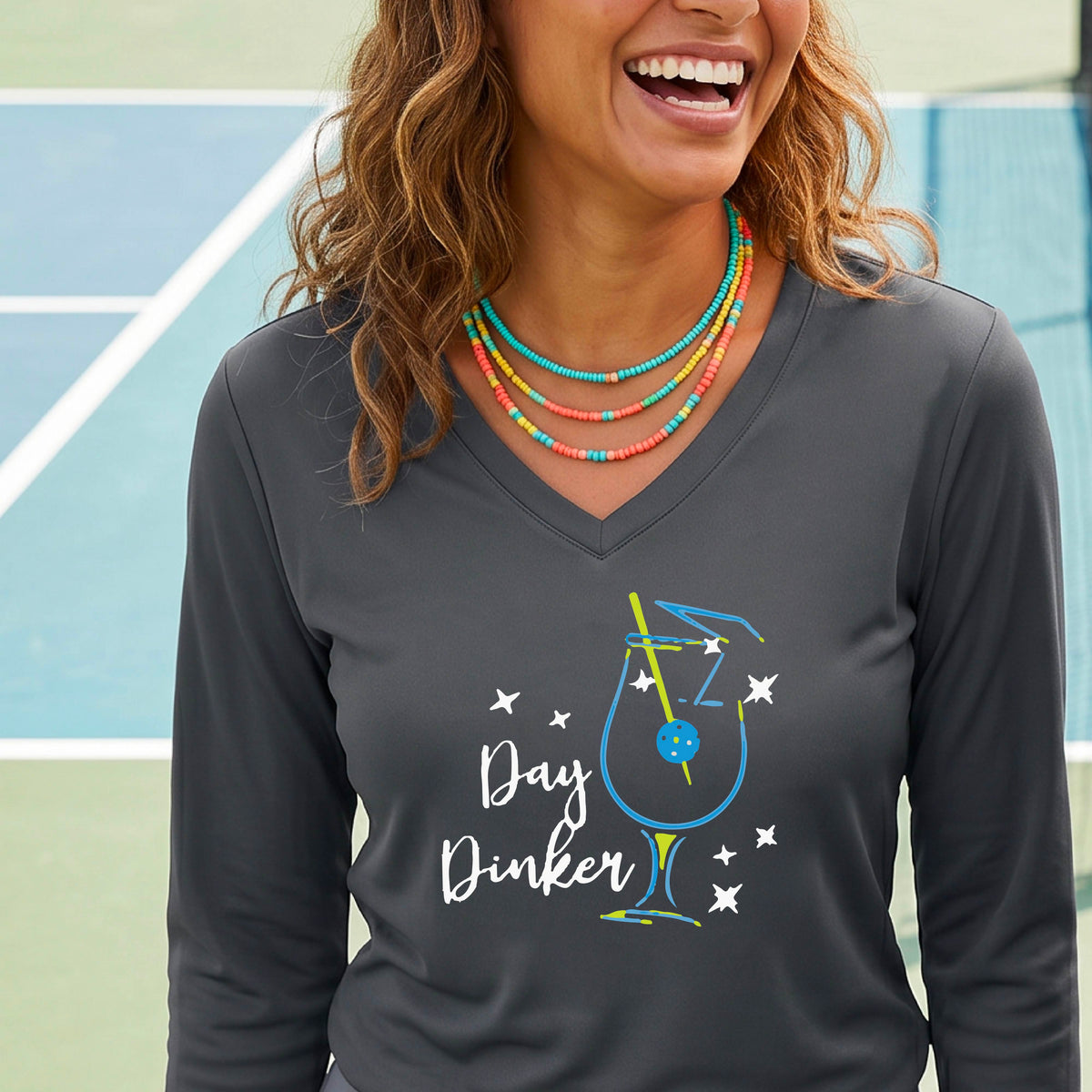 A woman wearing a dark gray long-sleeve V-neck t-shirt with a 'Day Dinker' cocktail martini graphic on the front, with a pickleball olive garnish.