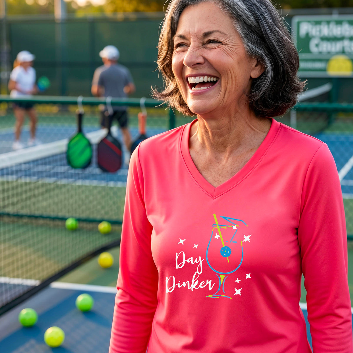 A woman wearing a coral pink long-sleeve V-neck t-shirt with a 'Day Dinker' cocktail martini graphic on the front, with a pickleball olive garnish.