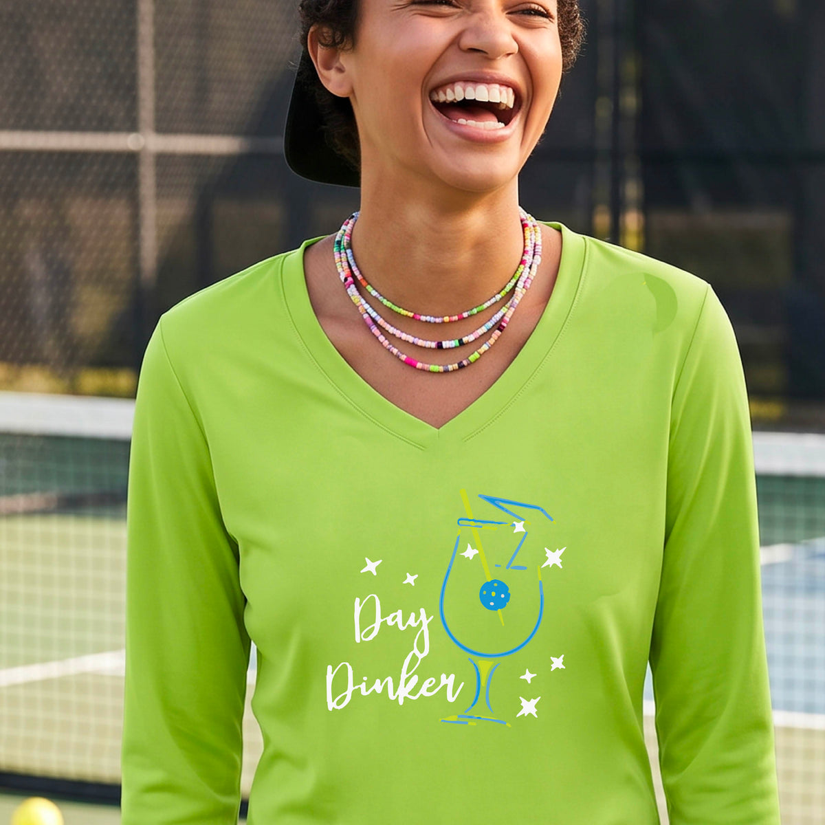 A woman wearing a lime green long-sleeve V-neck t-shirt with a 'Day Dinker' cocktail martini graphic on the front, with a pickleball olive garnish.