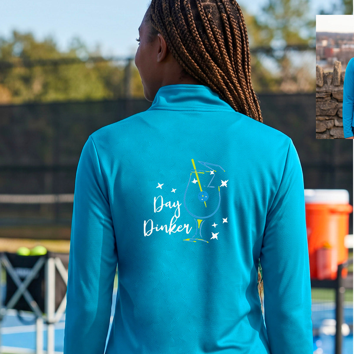 A woman wearing a light blue quarter-zip shirt with a martini cocktail graphic and a pickleball olive garnish, standing on a pickleball court. 