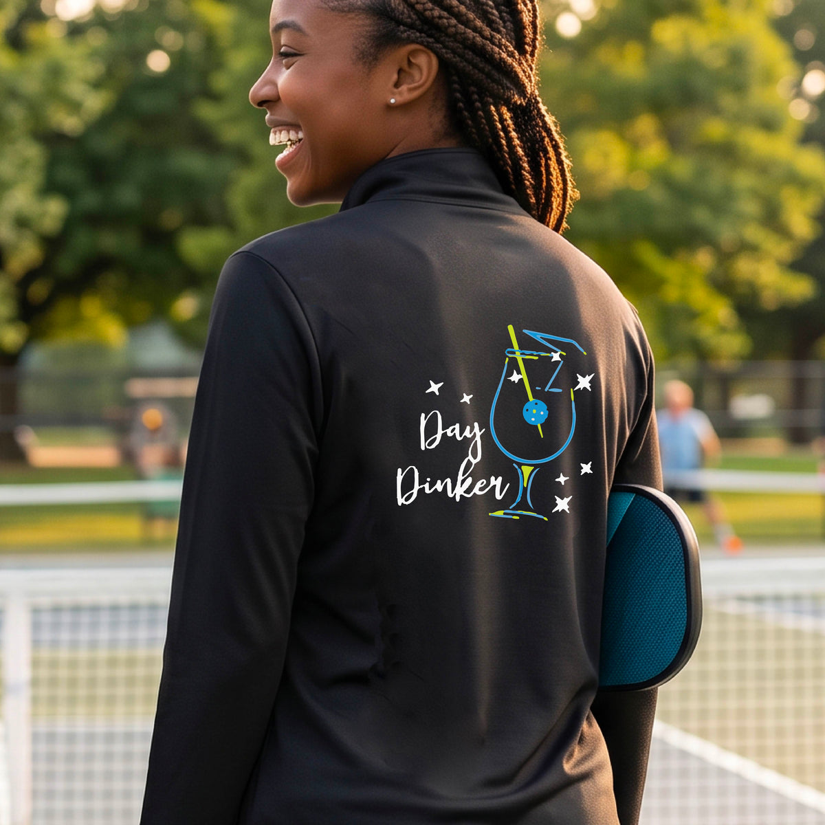 A woman wearing a black quarter-zip shirt with a martini cocktail graphic and a pickleball olive garnish, standing on a pickleball court. 