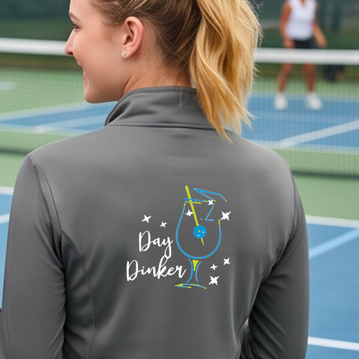 A woman wearing a gray quarter-zip shirt with a martini cocktail graphic and a pickleball olive garnish, standing on a pickleball court. 
