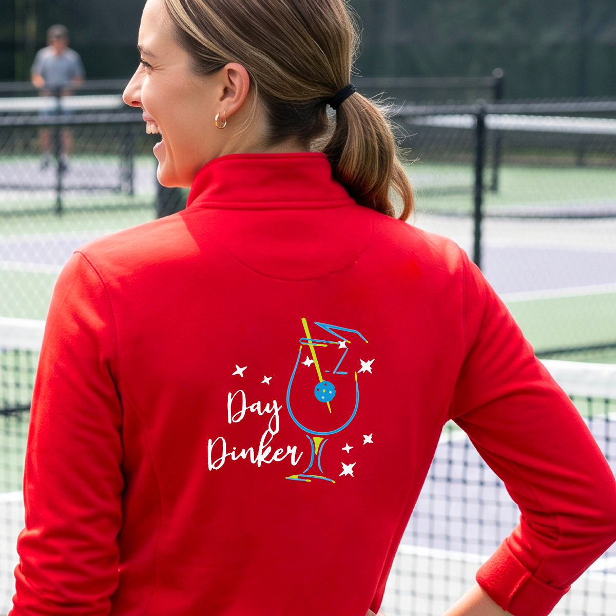 A woman wearing a red quarter-zip shirt with a martini cocktail graphic and a pickleball olive garnish, standing on a pickleball court. 
