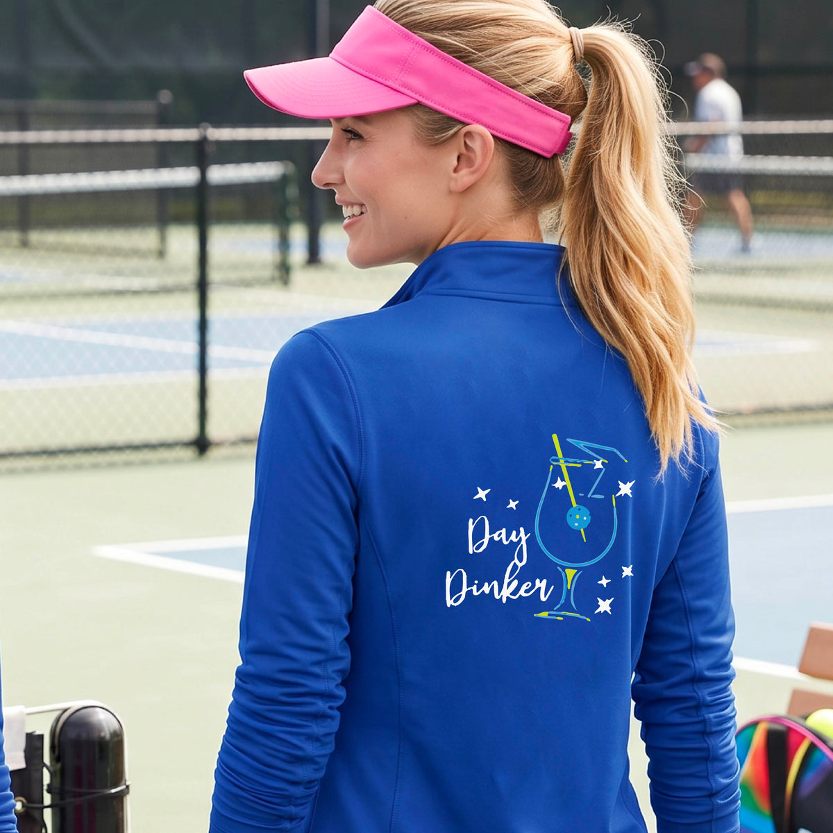 A woman wearing a royal blue quarter-zip shirt with a martini cocktail graphic and a pickleball olive garnish, standing on a pickleball court. 