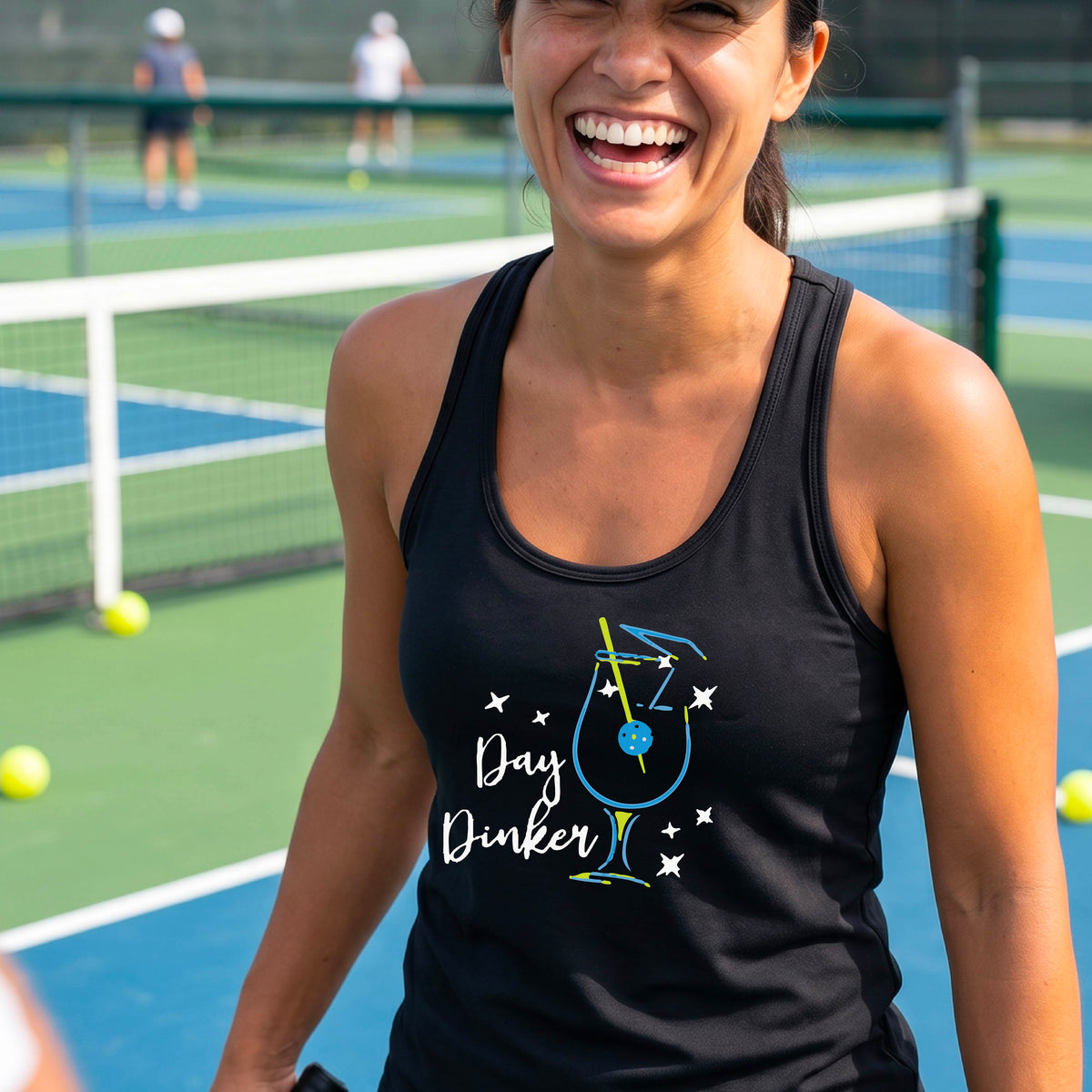 A woman wearing a black athletic tank top with a colorful 'Day Dinker' martini cocktail graphic with a pickleball olive as a garnish. on the front, standing on a pickleball court.