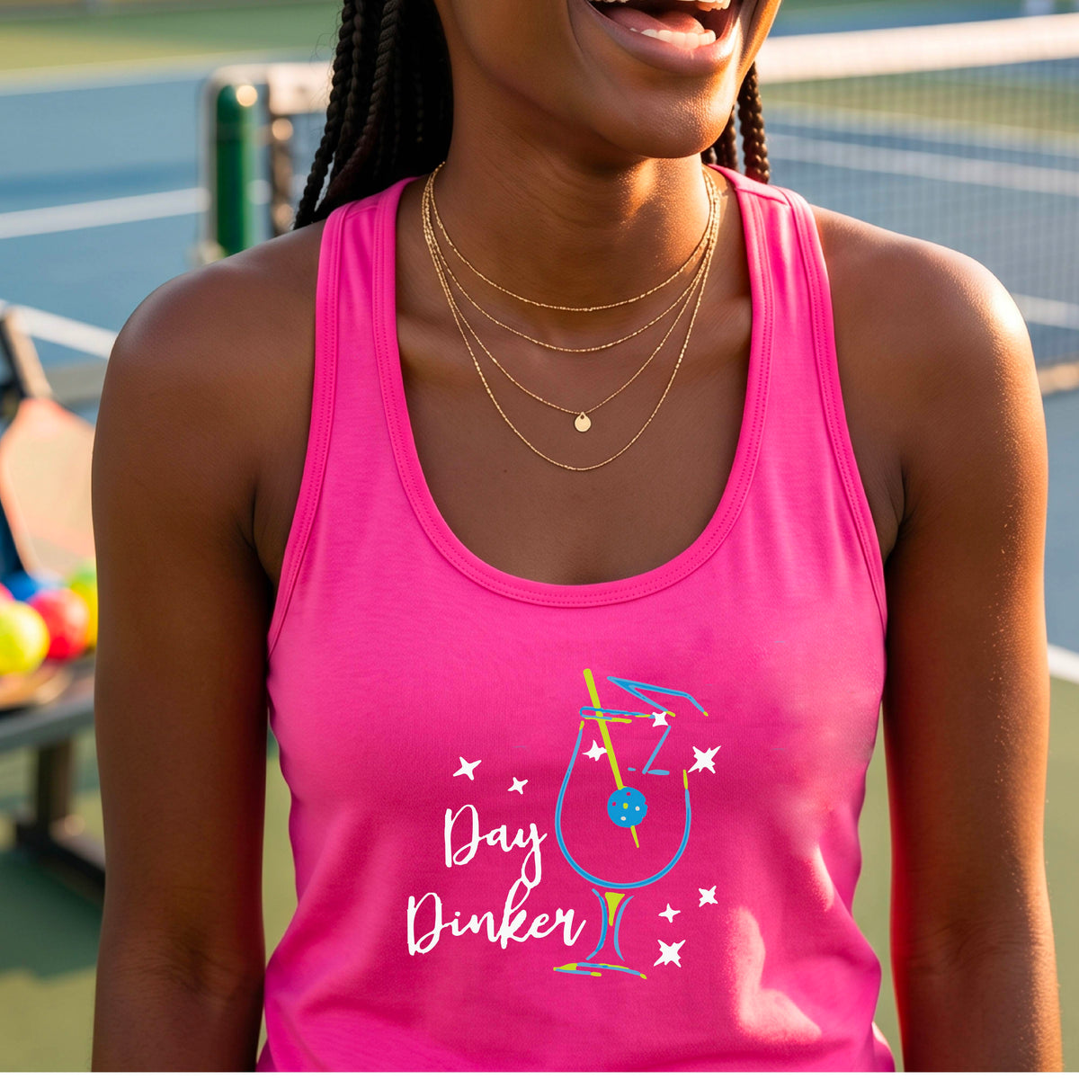 A woman wearing a bright pink athletic tank top with a colorful 'Day Dinker' martini cocktail graphic with a pickleball olive as a garnish. on the front, standing on a pickleball court.
