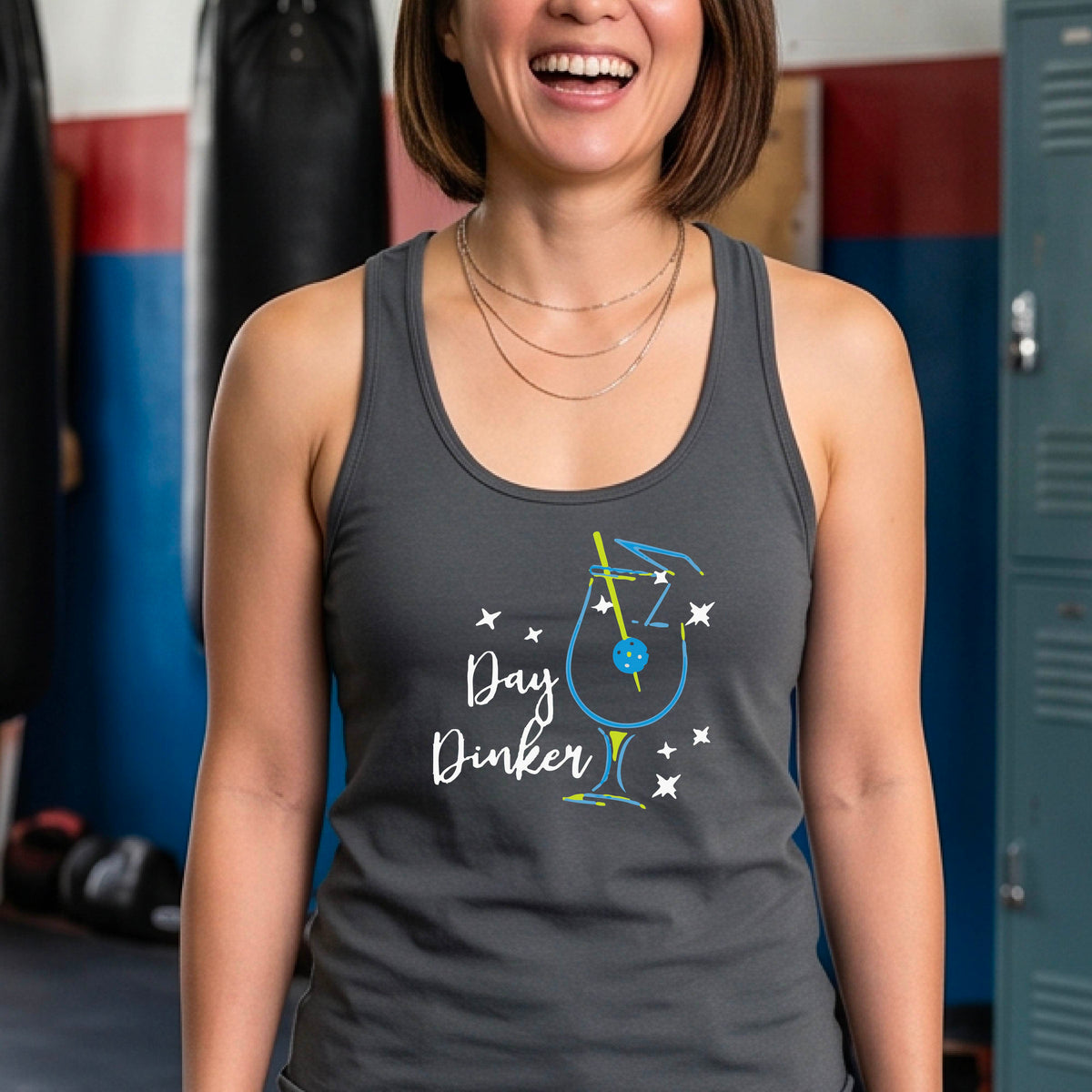 A woman wearing a dark gray athletic tank top with a colorful 'Day Dinker' martini cocktail graphic with a pickleball olive as a garnish. on the front, standing on a pickleball court.