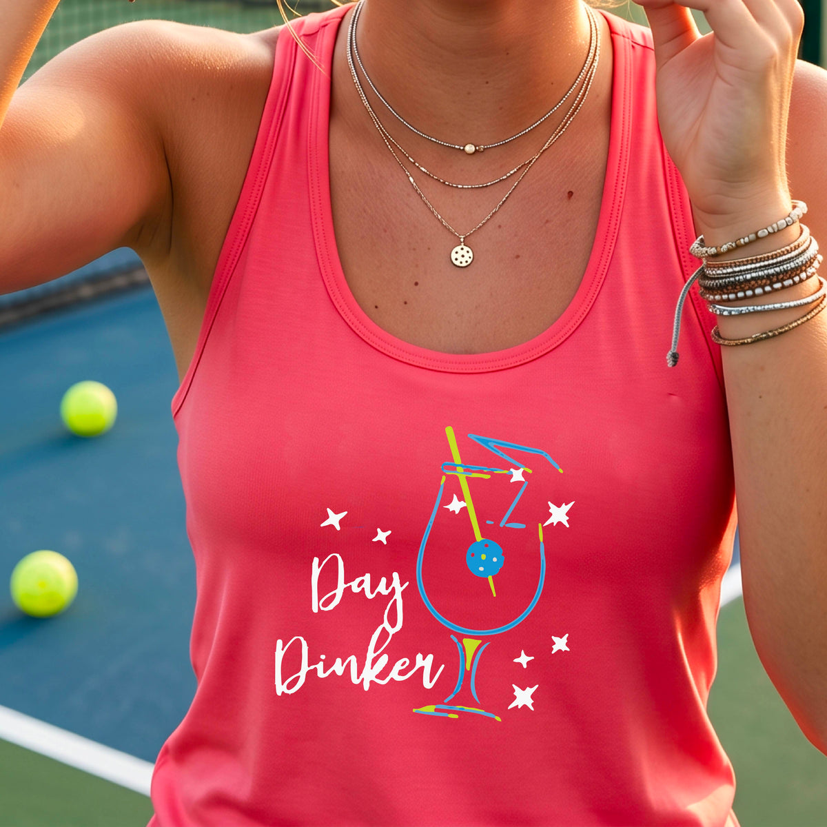 A woman wearing a coral pink athletic tank top with a colorful 'Day Dinker' martini cocktail graphic with a pickleball olive as a garnish. on the front, standing on a pickleball court.