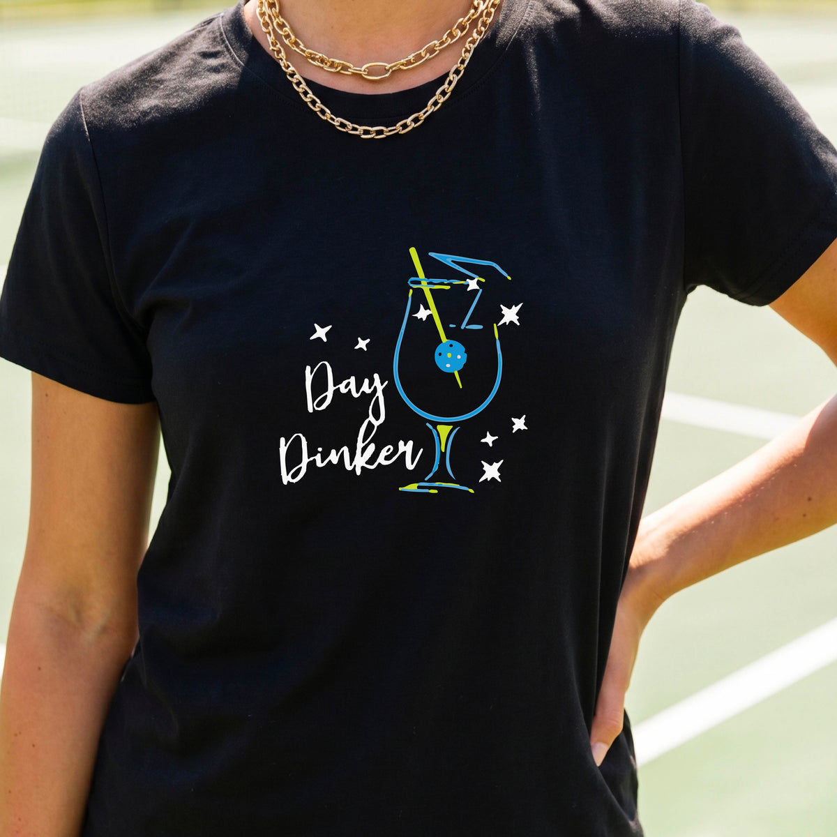 A person wearing a black short-sleeve T-shirt with a colorful graphic of a cocktail martini glass and the words 'Day Dinker' on the front, with a pickleball olive garnish, photographed outdoors on a pickleball court.