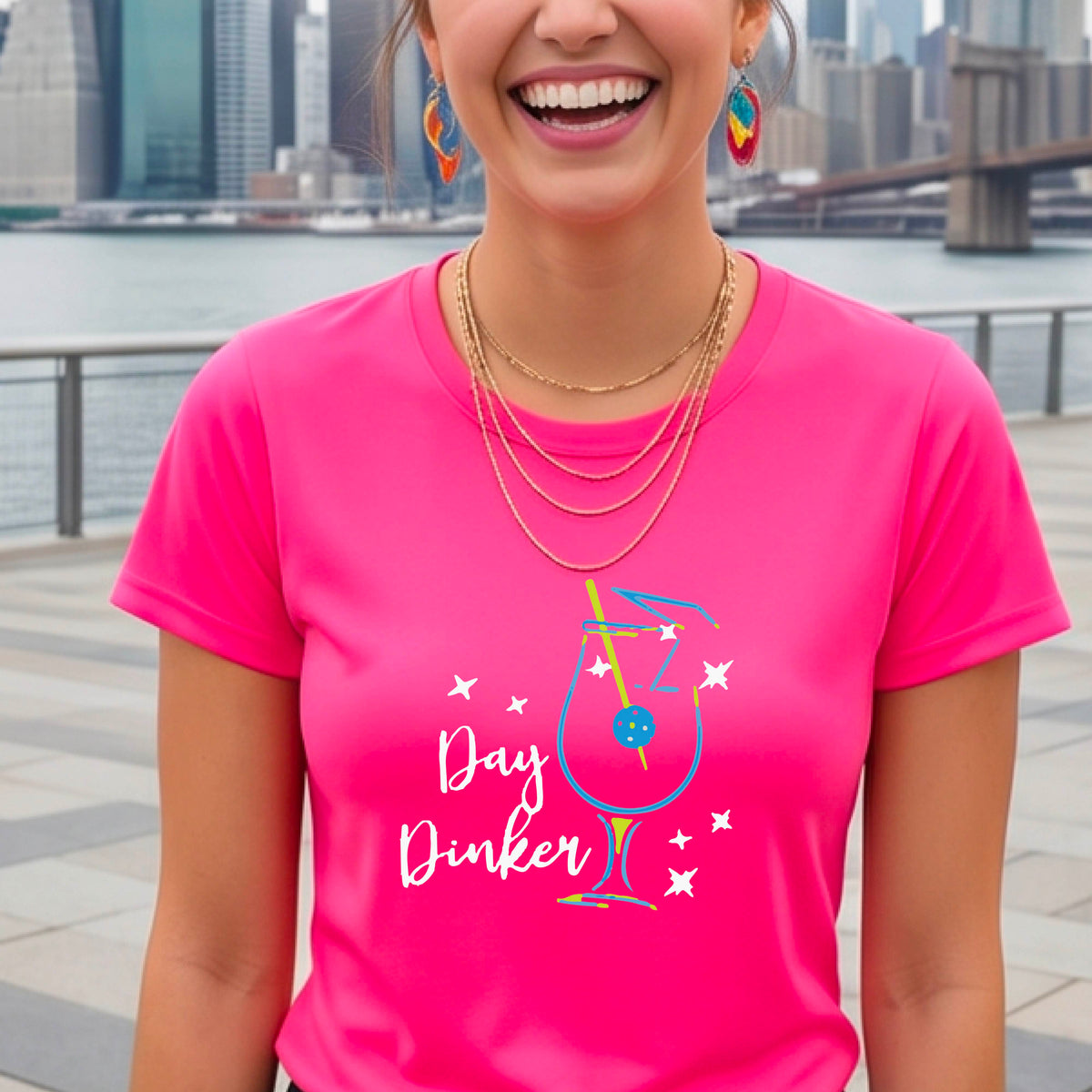 A person wearing a pink short-sleeve T-shirt with a colorful graphic of a cocktail martini glass and the words 'Day Dinker' on the front, with a pickleball olive garnish.