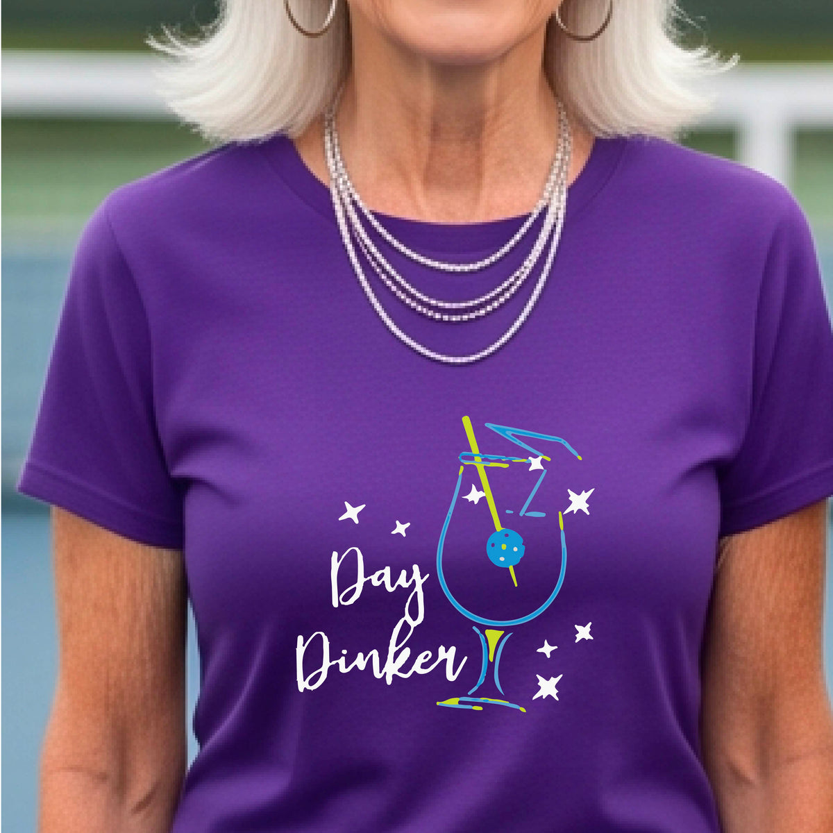 A person wearing a purple short-sleeve T-shirt with a colorful graphic of a cocktail martini glass and the words 'Day Dinker' on the front, with a pickleball olive garnish, photographed outdoors on a pickleball court.