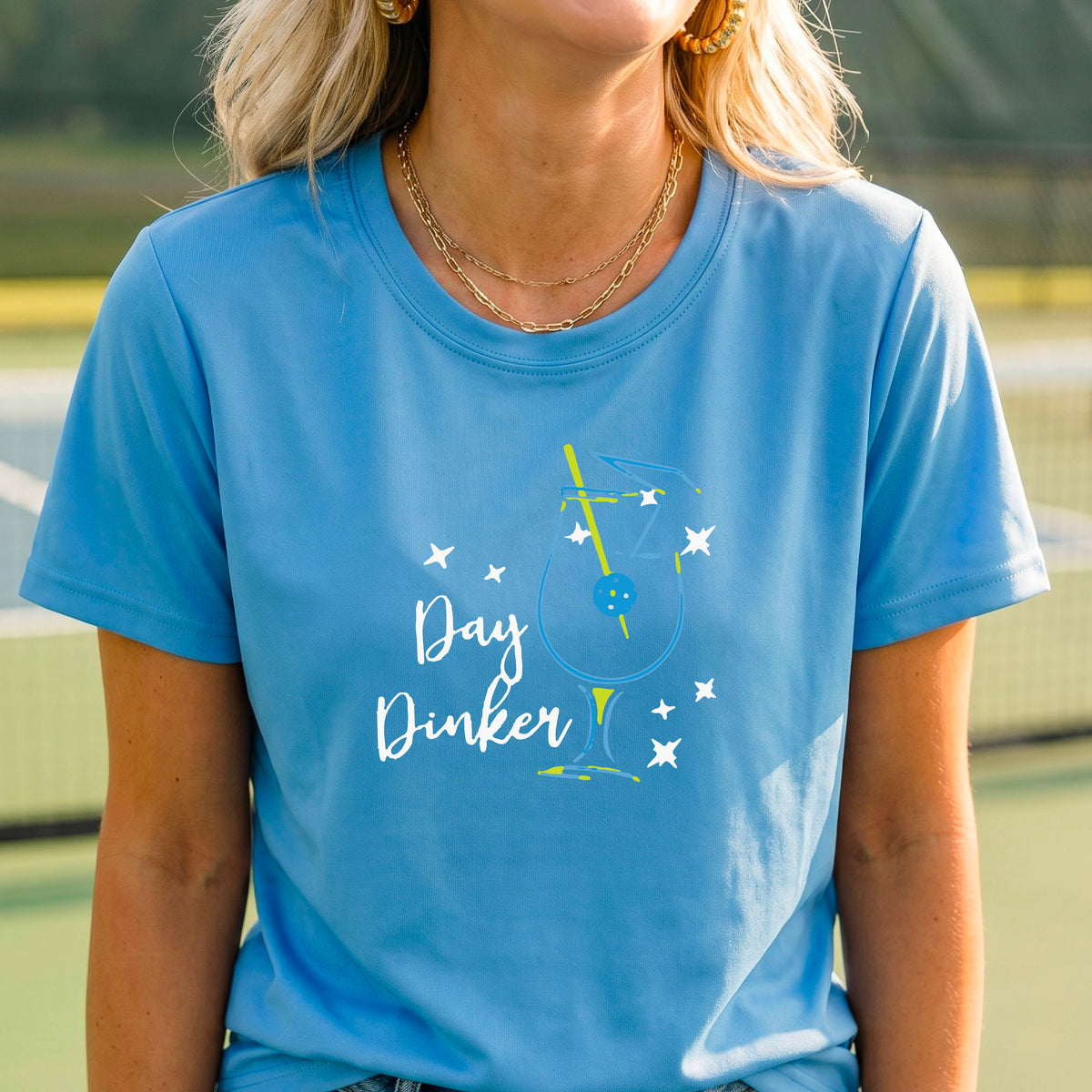 A person wearing a light blue short-sleeve T-shirt with a colorful graphic of a cocktail martini glass and the words 'Day Dinker' on the front, with a pickleball olive garnish, photographed outdoors on a pickleball court.