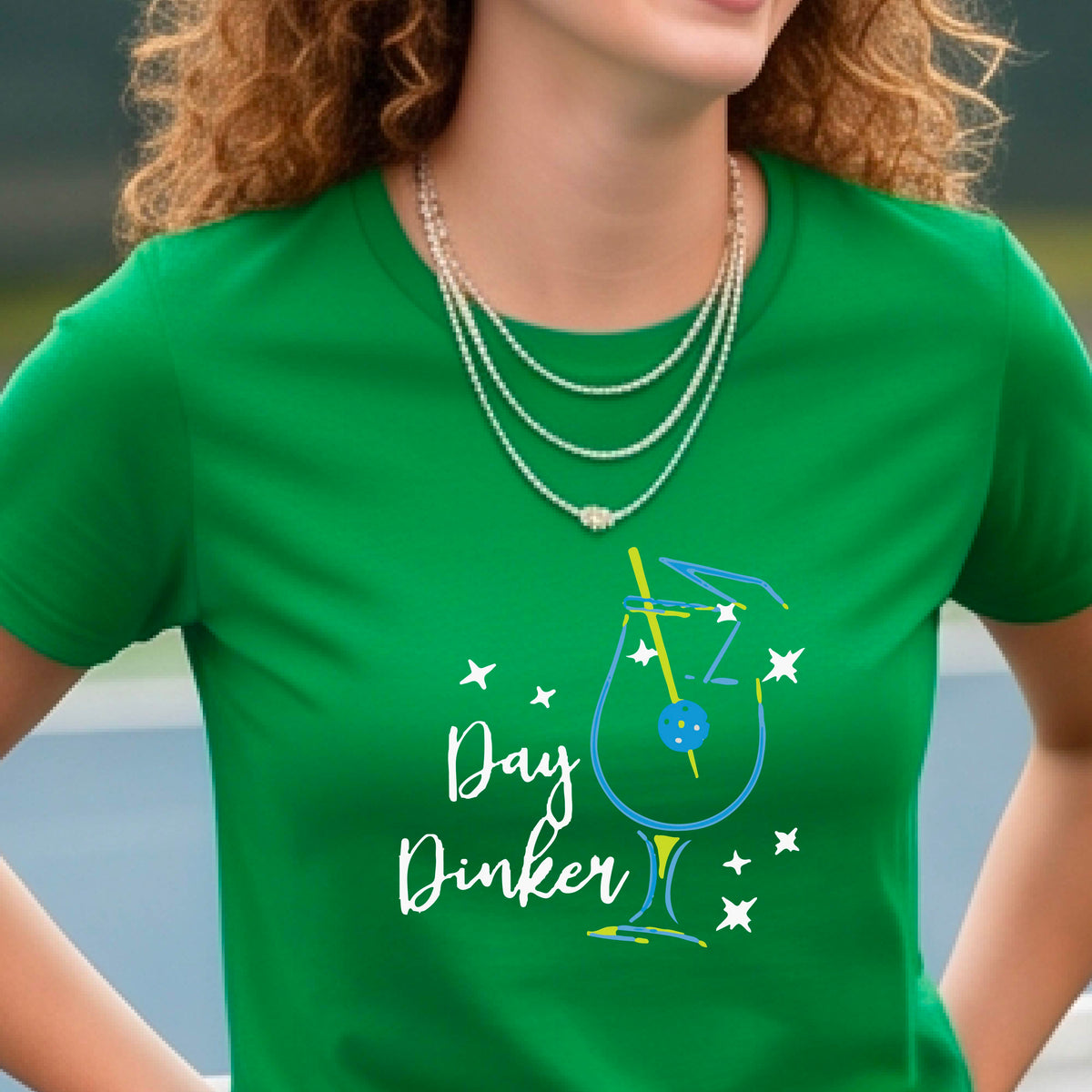 A person wearing a green short-sleeve T-shirt with a colorful graphic of a cocktail martini glass and the words 'Day Dinker' on the front, with a pickleball olive garnish, photographed outdoors on a pickleball court.