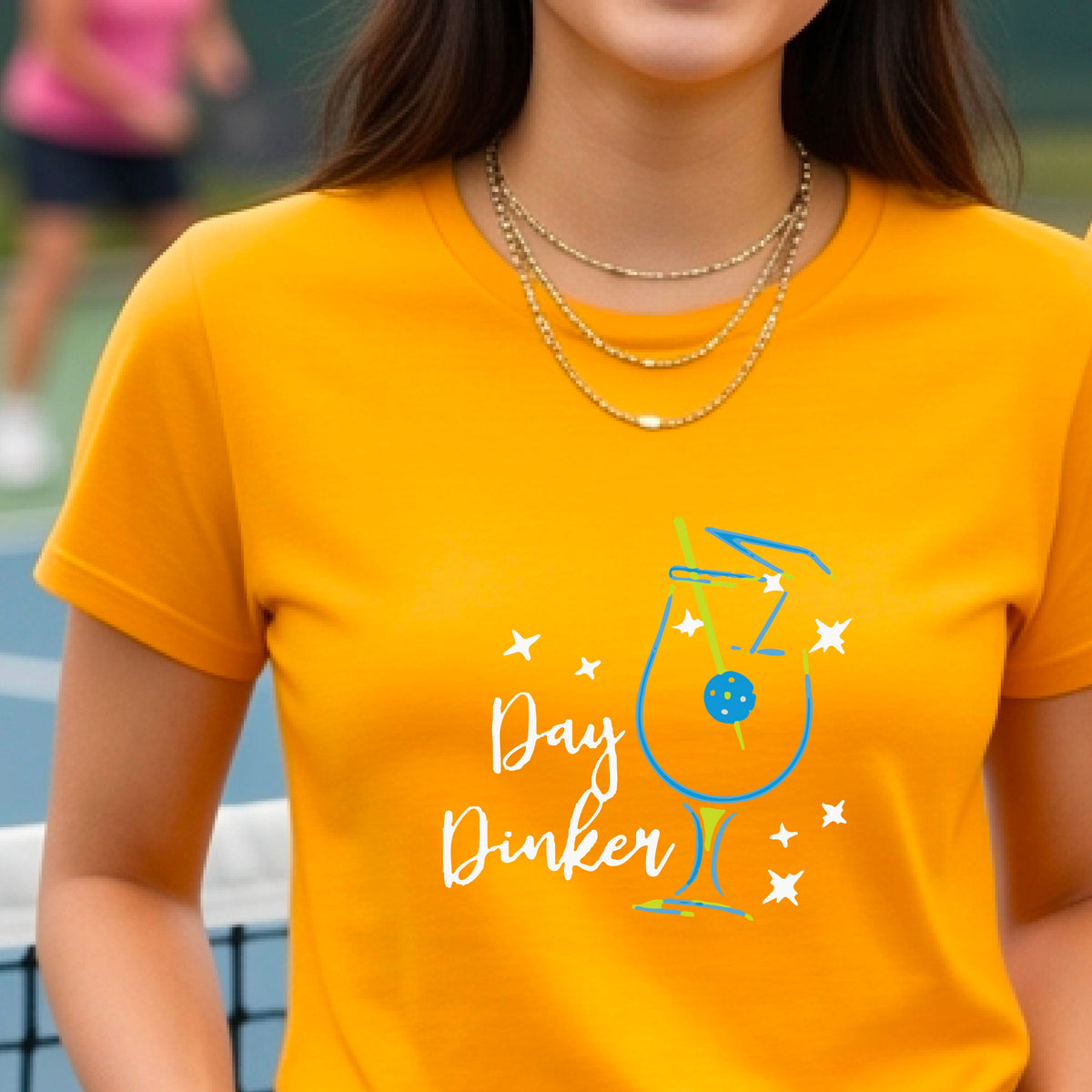 A person wearing a mustard yellow gold short-sleeve T-shirt with a colorful graphic of a cocktail martini glass and the words 'Day Dinker' on the front, with a pickleball olive garnish, photographed outdoors on a pickleball court.