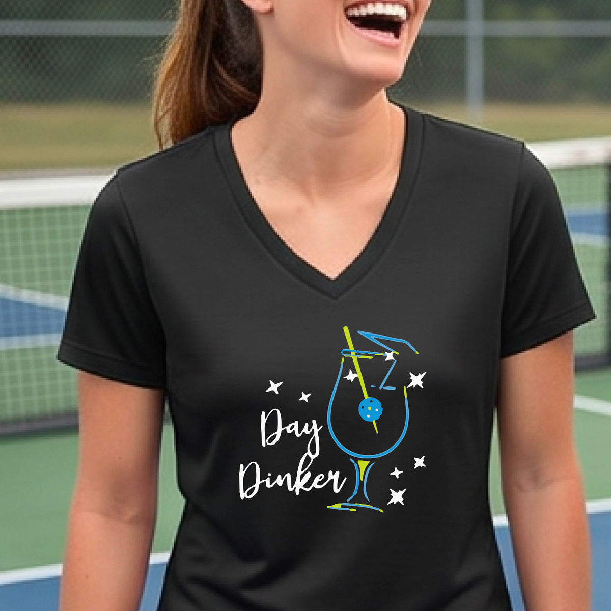A woman wearing a black V-neck t-shirt with a colorful 'Day Dinker' martinin cocktail graphic with a pickleball garnish on the front.