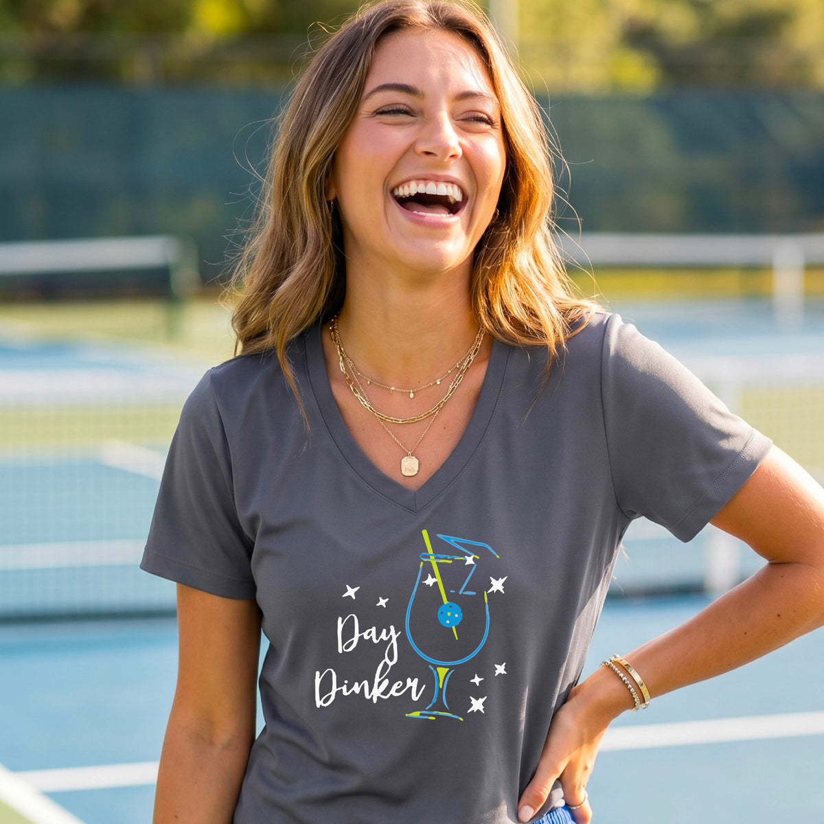 A woman wearing a dark gray V-neck t-shirt with a colorful 'Day Dinker' martinin cocktail graphic with a pickleball garnish on the front.