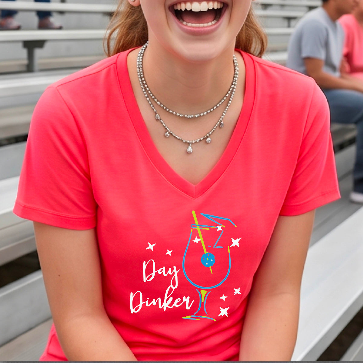 A woman wearing a bright coral pink V-neck t-shirt with a colorful 'Day Dinker' martinin cocktail graphic with a pickleball garnish on the front.