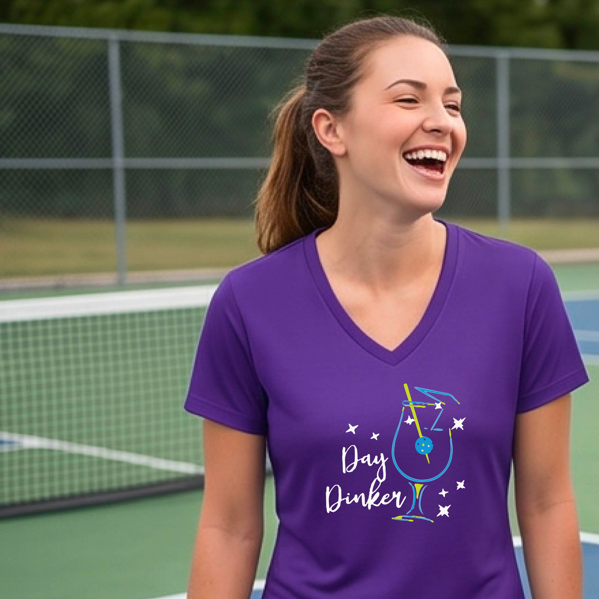 A woman wearing a purple V-neck t-shirt with a colorful 'Day Dinker' martinin cocktail graphic with a pickleball garnish on the front.