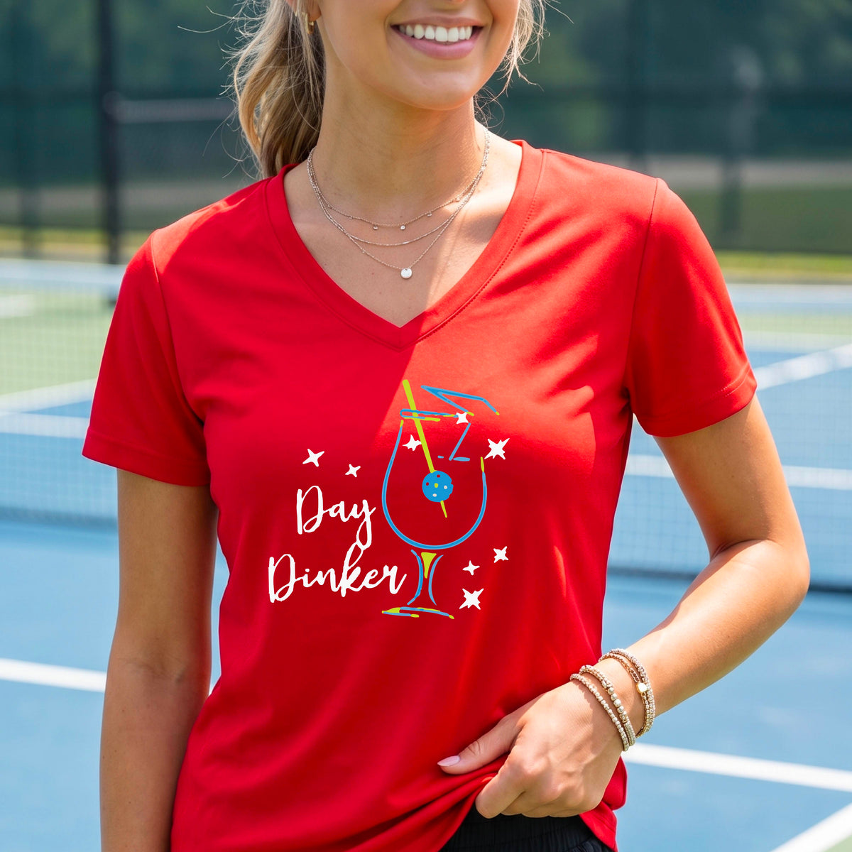 A woman wearing a bright red V-neck t-shirt with a colorful 'Day Dinker' martinin cocktail graphic with a pickleball garnish on the front.
