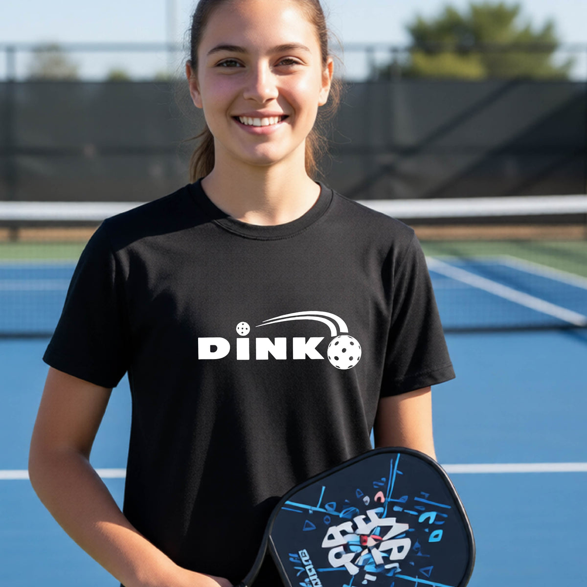 A young person wearing a black short-sleeve t-shirt with a pickleball design of text 'DINK' with a white pickleall in flight, standing on a pickleball court.