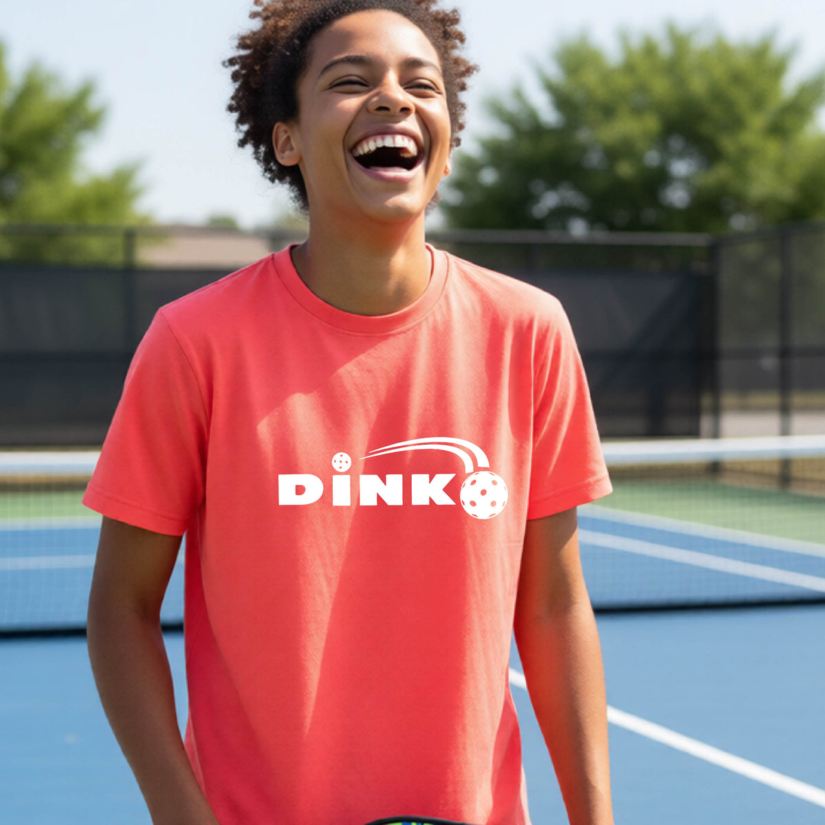 A young person wearing a coral pink short-sleeve t-shirt with a pickleball design of text 'DINK' with a white pickleall in flight, standing on a pickleball court.