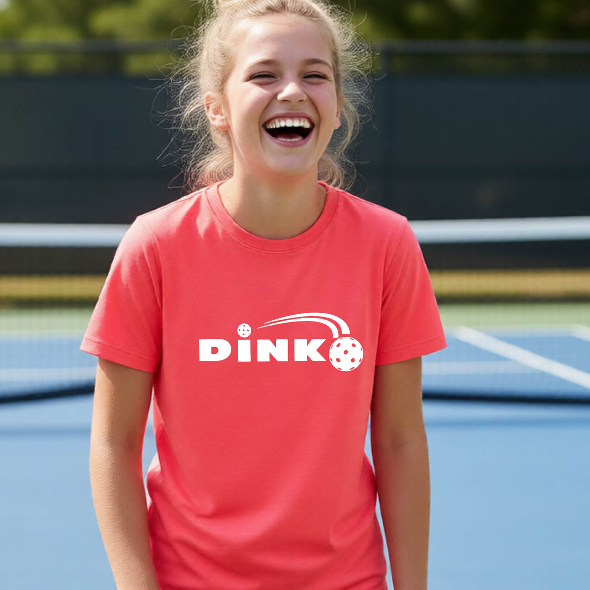 A young person wearing a coral pink short-sleeve t-shirt with a pickleball design of text 'DINK' with a white pickleall in flight, standing on a pickleball court.
