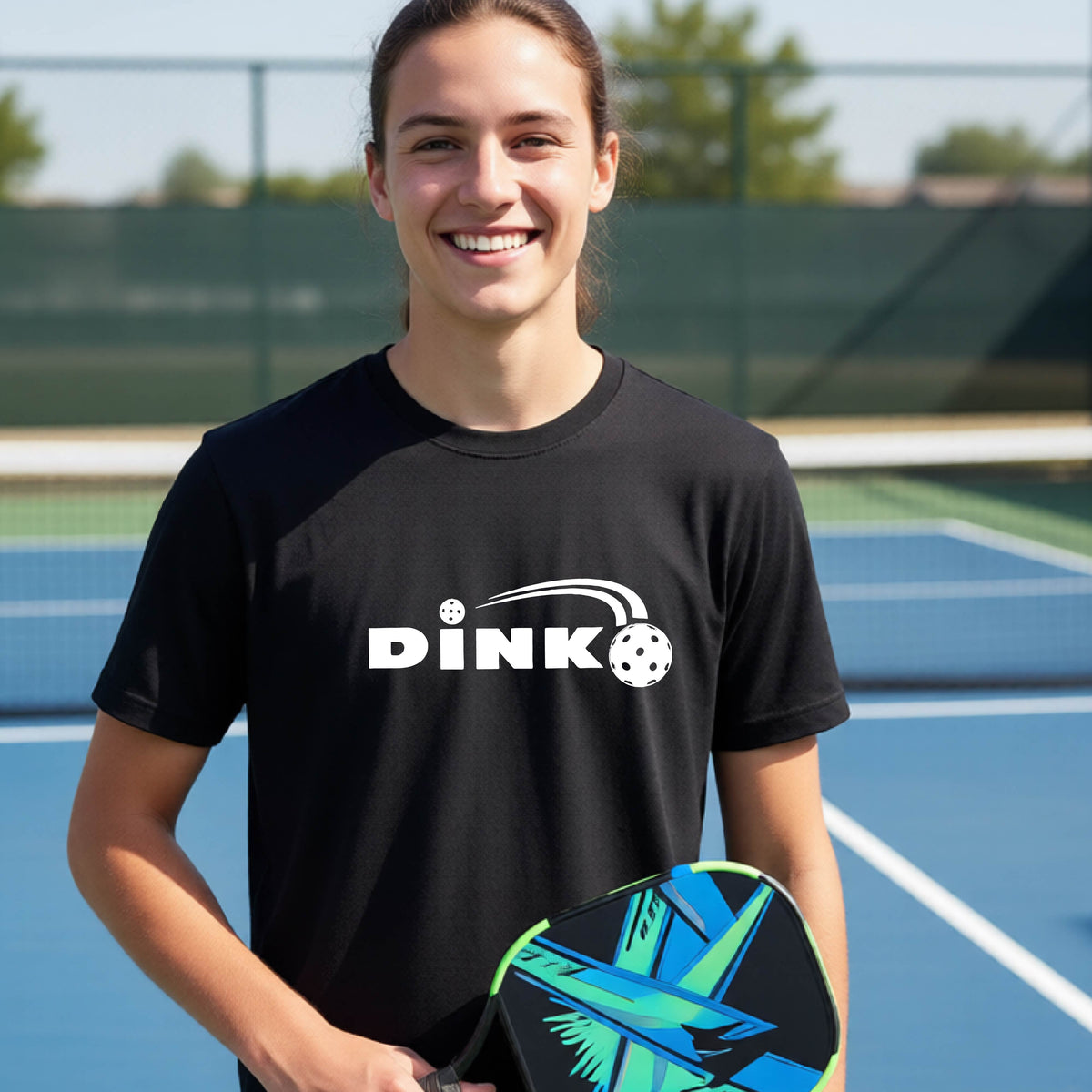 A young person wearing a black short-sleeve t-shirt with a pickleball design of text 'DINK' with a white pickleall in flight, standing on a pickleball court.