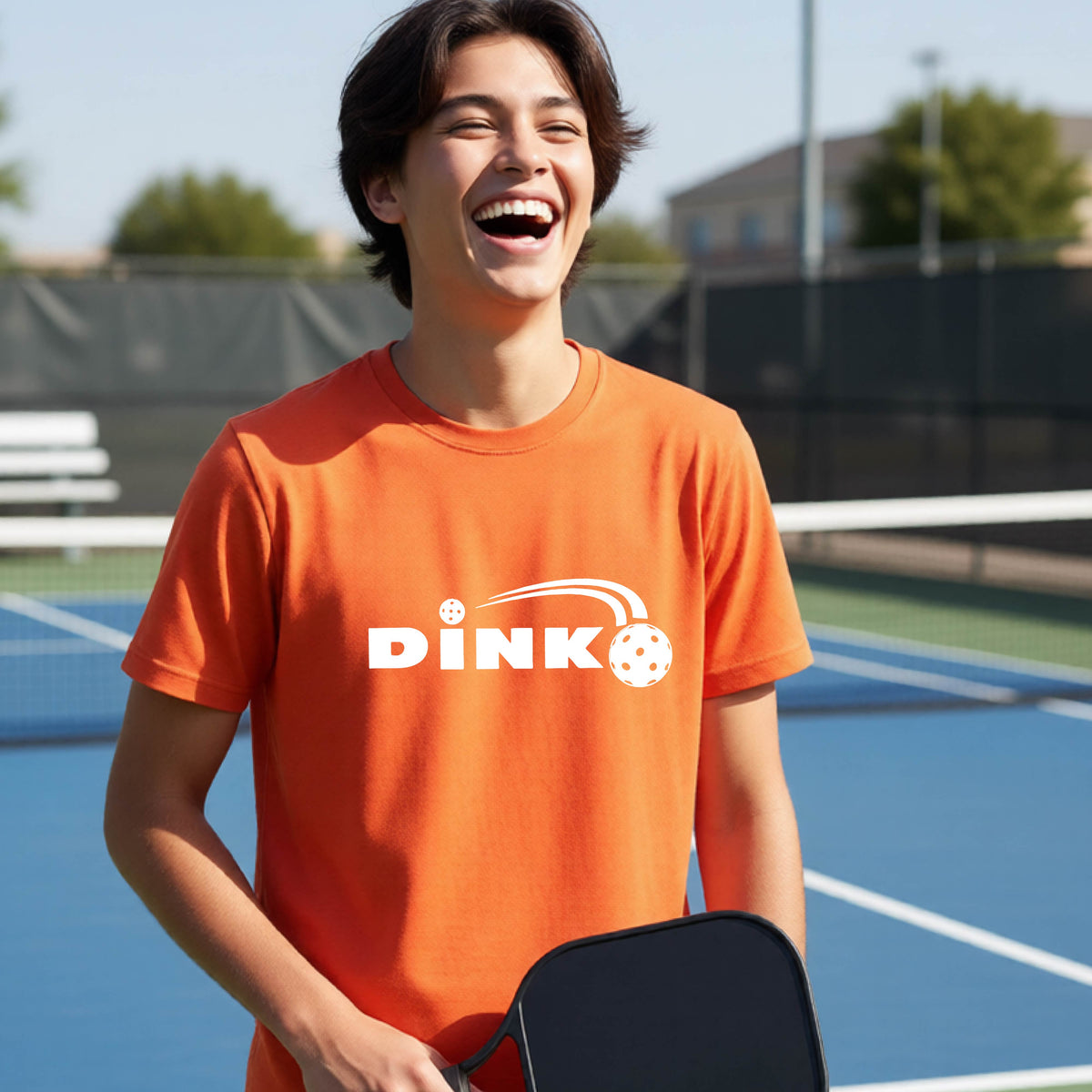 A young person wearing an orange short-sleeve t-shirt with a pickleball design of text 'DINK' with a white pickleall in flight, standing on a pickleball court.