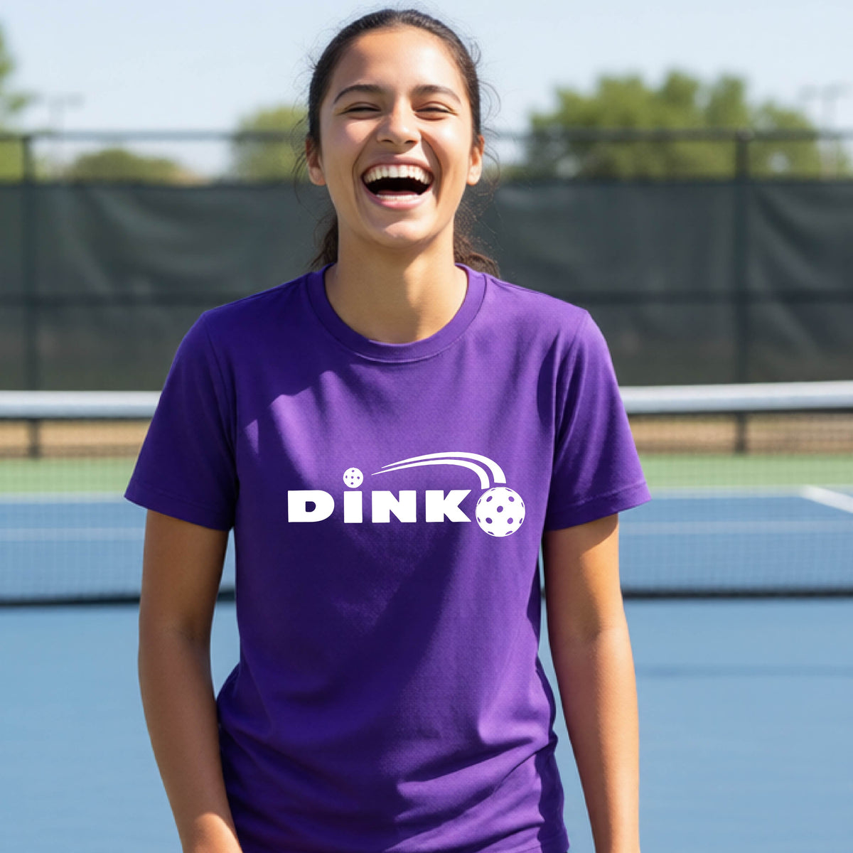 A young person wearing a purple short-sleeve t-shirt with a pickleball design of text 'DINK' with a white pickleall in flight, standing on a pickleball court.