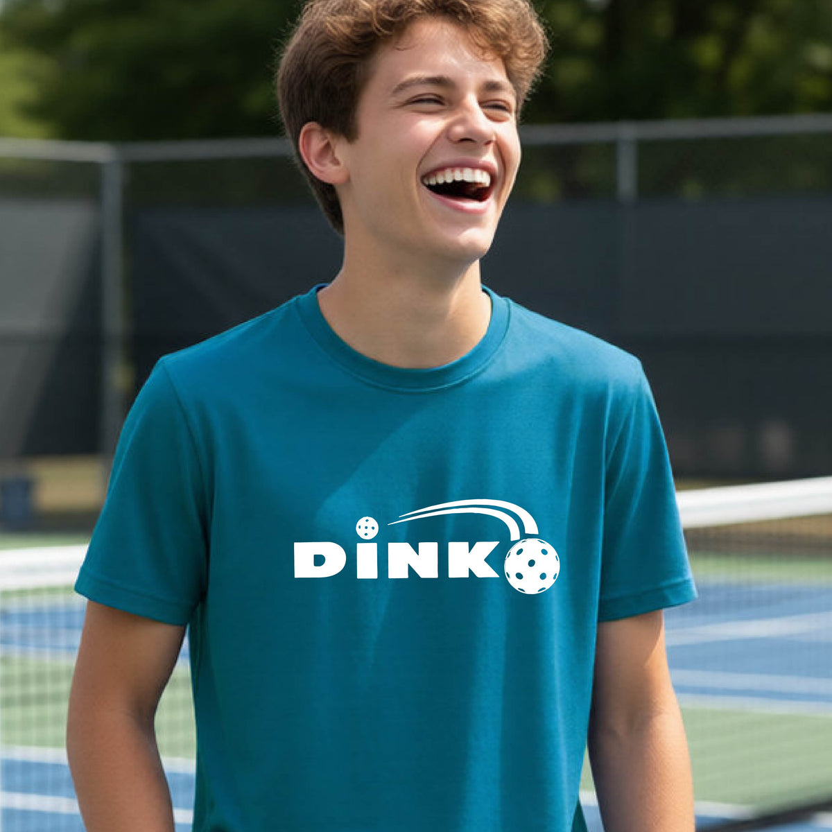A young person wearing a teal short-sleeve t-shirt with a pickleball design of text 'DINK' with a white pickleall in flight, standing on a pickleball court.