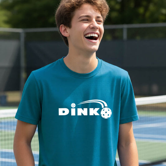 A young person wearing a teal short-sleeve t-shirt with a pickleball design of text 'DINK' with a white pickleall in flight, standing on a pickleball court.