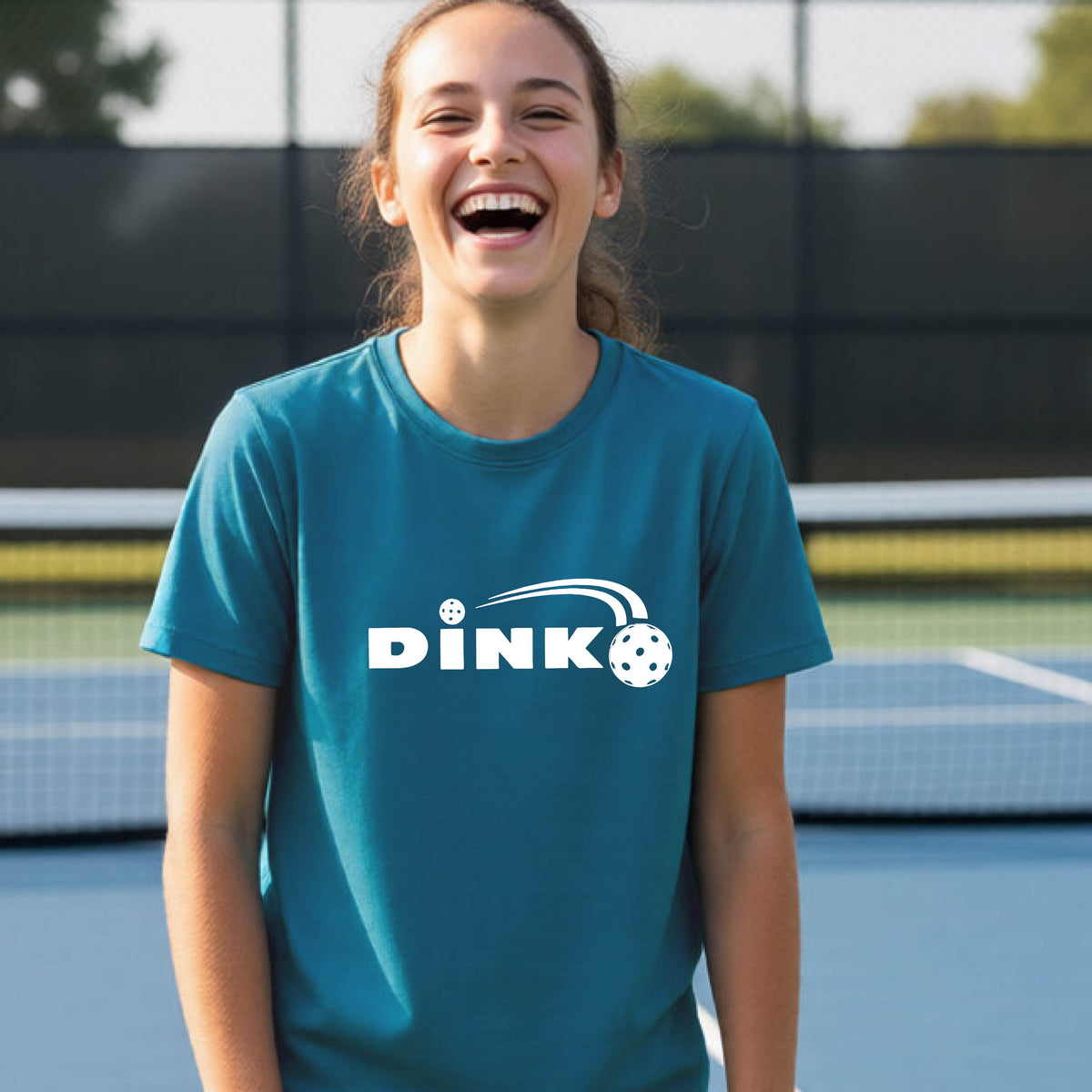 A young person wearing a teal short-sleeve t-shirt with a pickleball design of text 'DINK' with a white pickleall in flight, standing on a pickleball court.